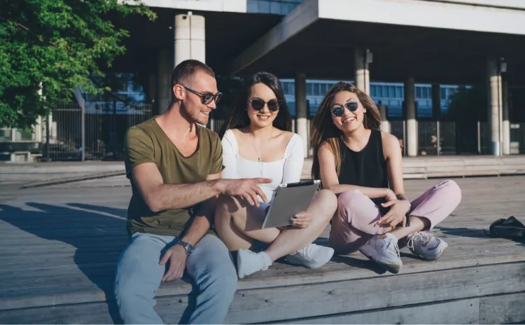 Internationale Studenten aus europäischen, arabischen und asiatischen Ländern, die auf einer Holzbank im freien mit einem Tablet lernen.